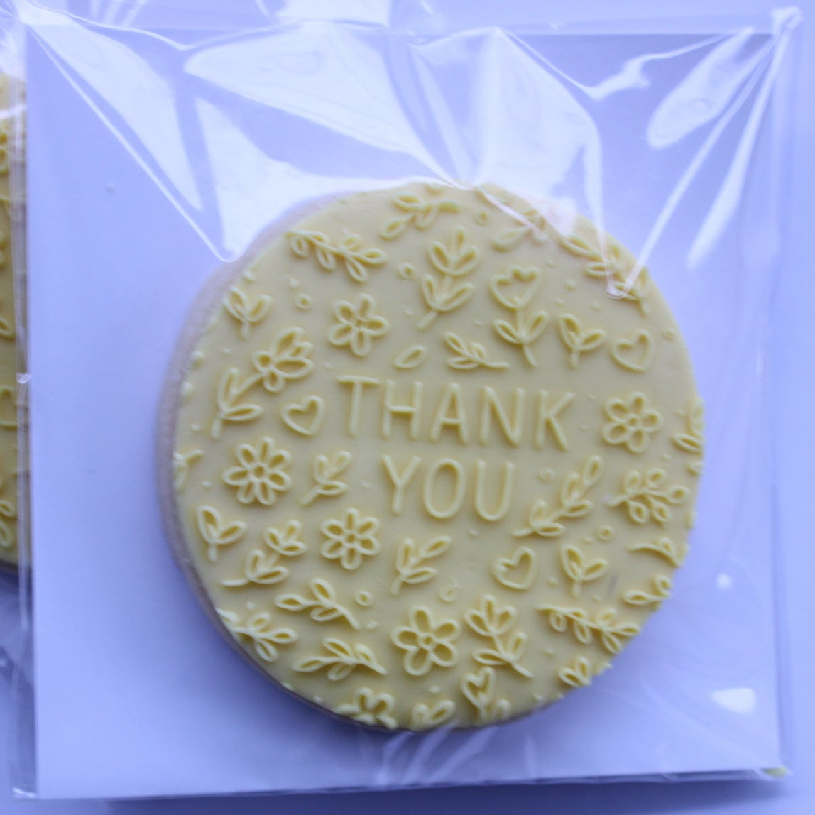 A round pale yellow decorative cookie stamped with floral patterns and the words 'THANK YOU' in the center, sealed in a clear plastic bag on a light lavender background.