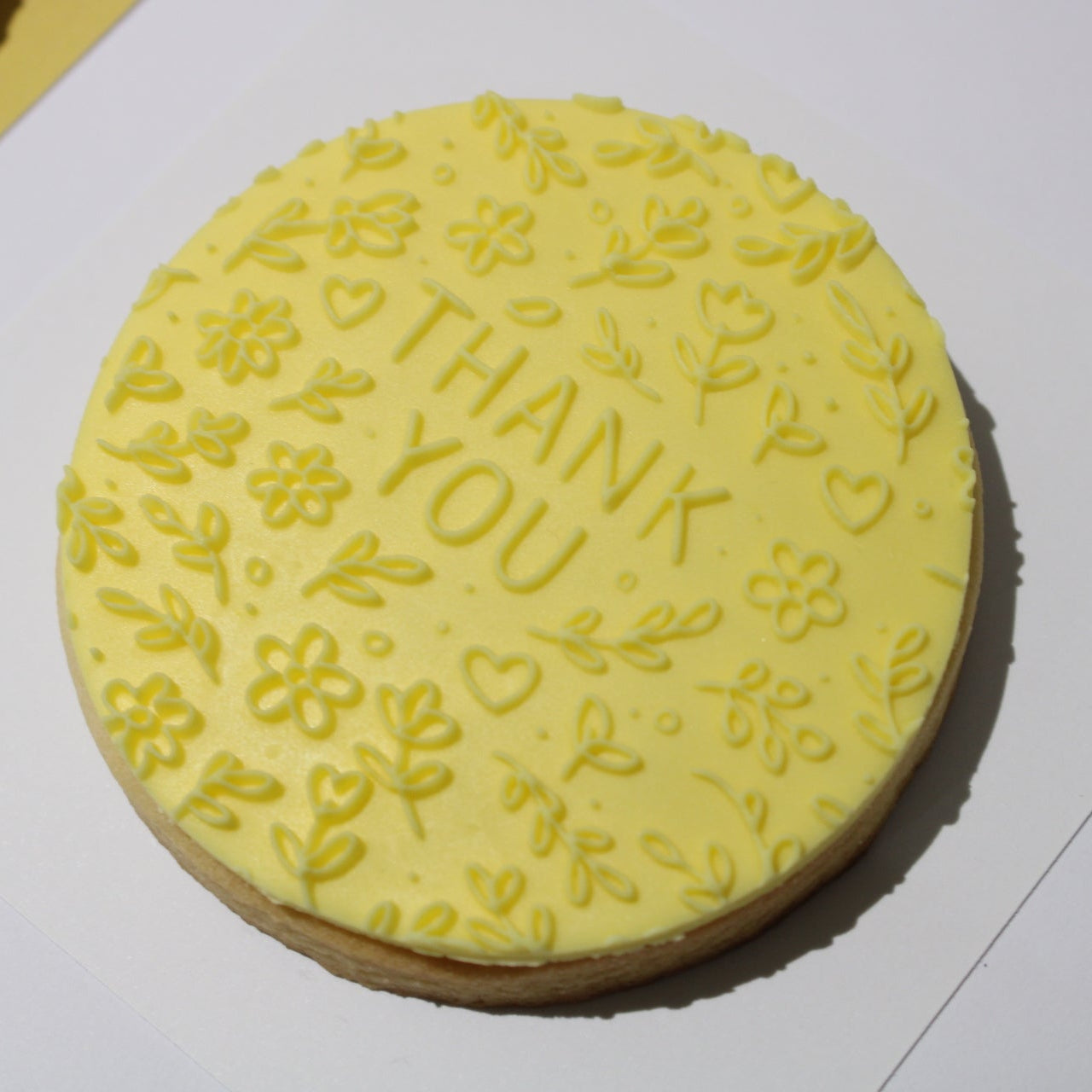 A round yellow frosted cookie with 'THANK YOU' and floral designs embossed on top, resting on a white surface.